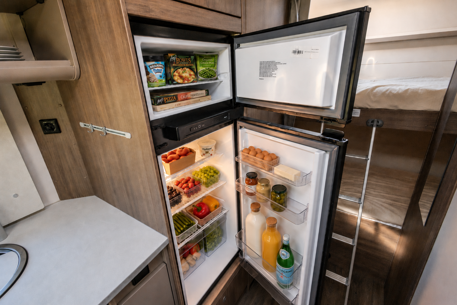 Van interior with open fridge and freezer, stocked shelves, adjacent counter, partial view of sleeping area and ladder.