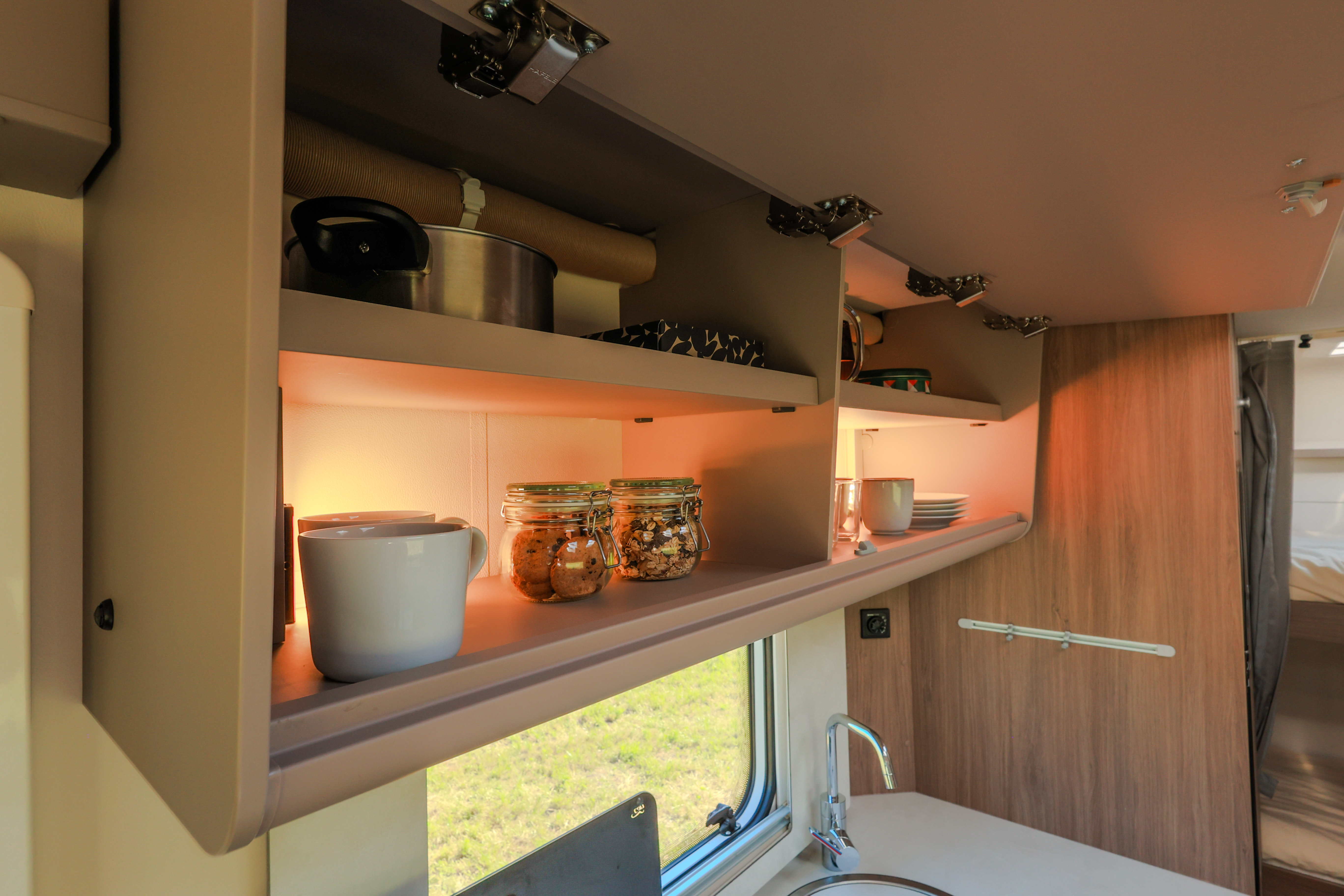 Overhead kitchen cabinets with dishes, jars, and cookware above a countertop and sink inside the van.