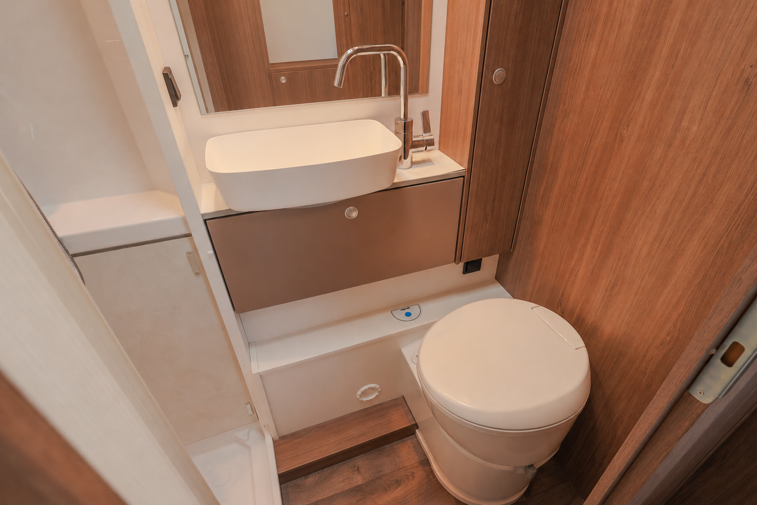 Compact van bathroom interior with toilet, sink, wooden cabinets, mirror, and storage shelves visible.