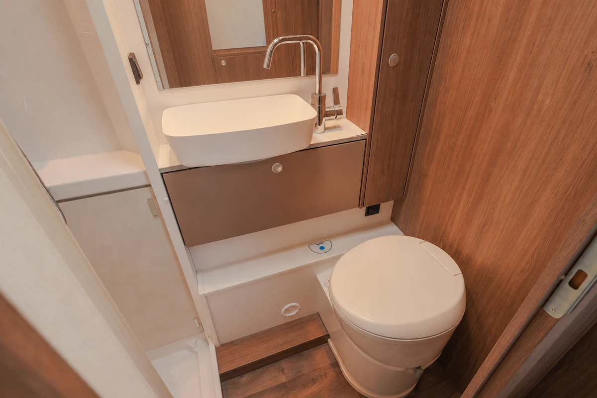 Compact van bathroom interior with toilet, sink, wooden cabinets, mirror, and storage shelves visible.