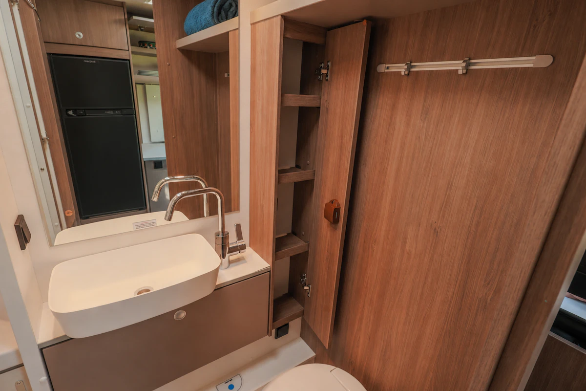 Van bathroom interior with sink, toilet, mirror, storage shelves, and wooden cabinets visible.