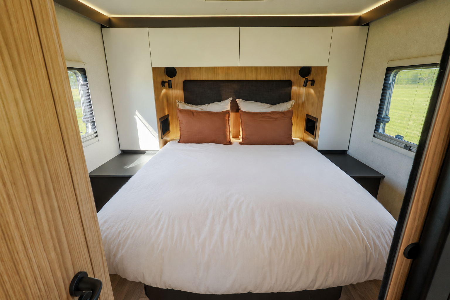 Van interior with large bed, overhead cabinets, side tables, windows on both sides, and wood panel accents.