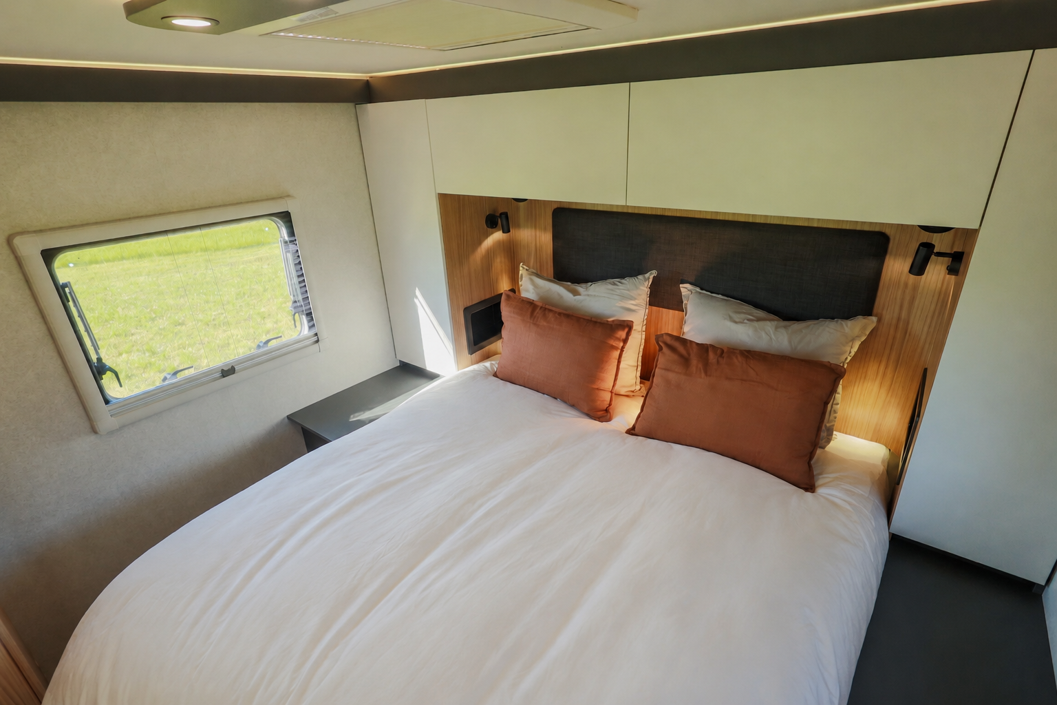 Van interior with double bed, overhead cabinets, two side tables, and a window on the left.