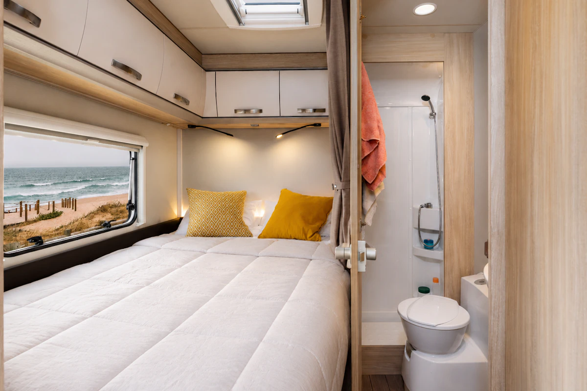 Van interior with double bed, overhead cabinets, window, ensuite bathroom with shower and toilet, modern light wood finish.