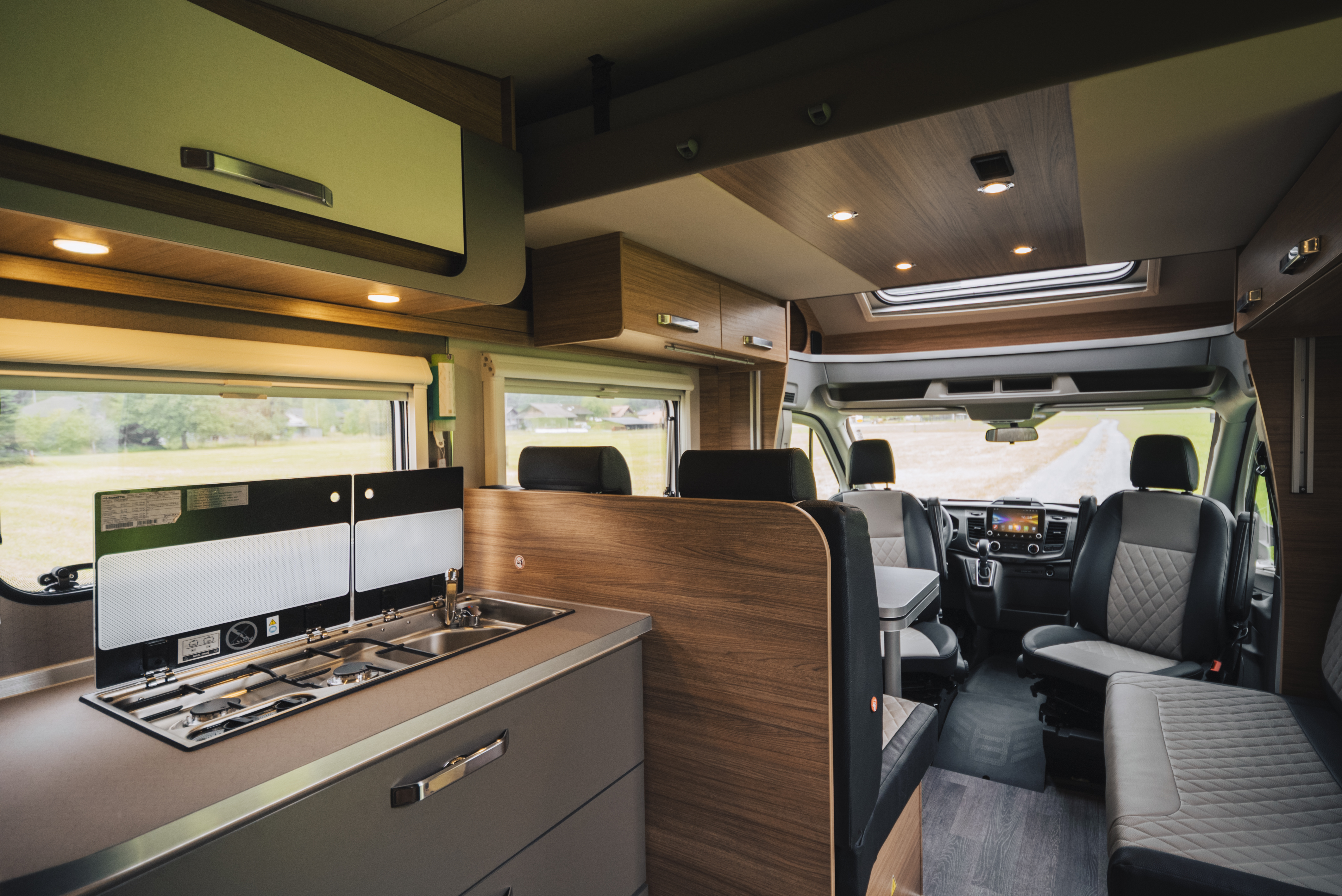 Camper van interior with kitchen, sink, stove, seating area, storage cabinets, and front driver and passenger seats visible.
