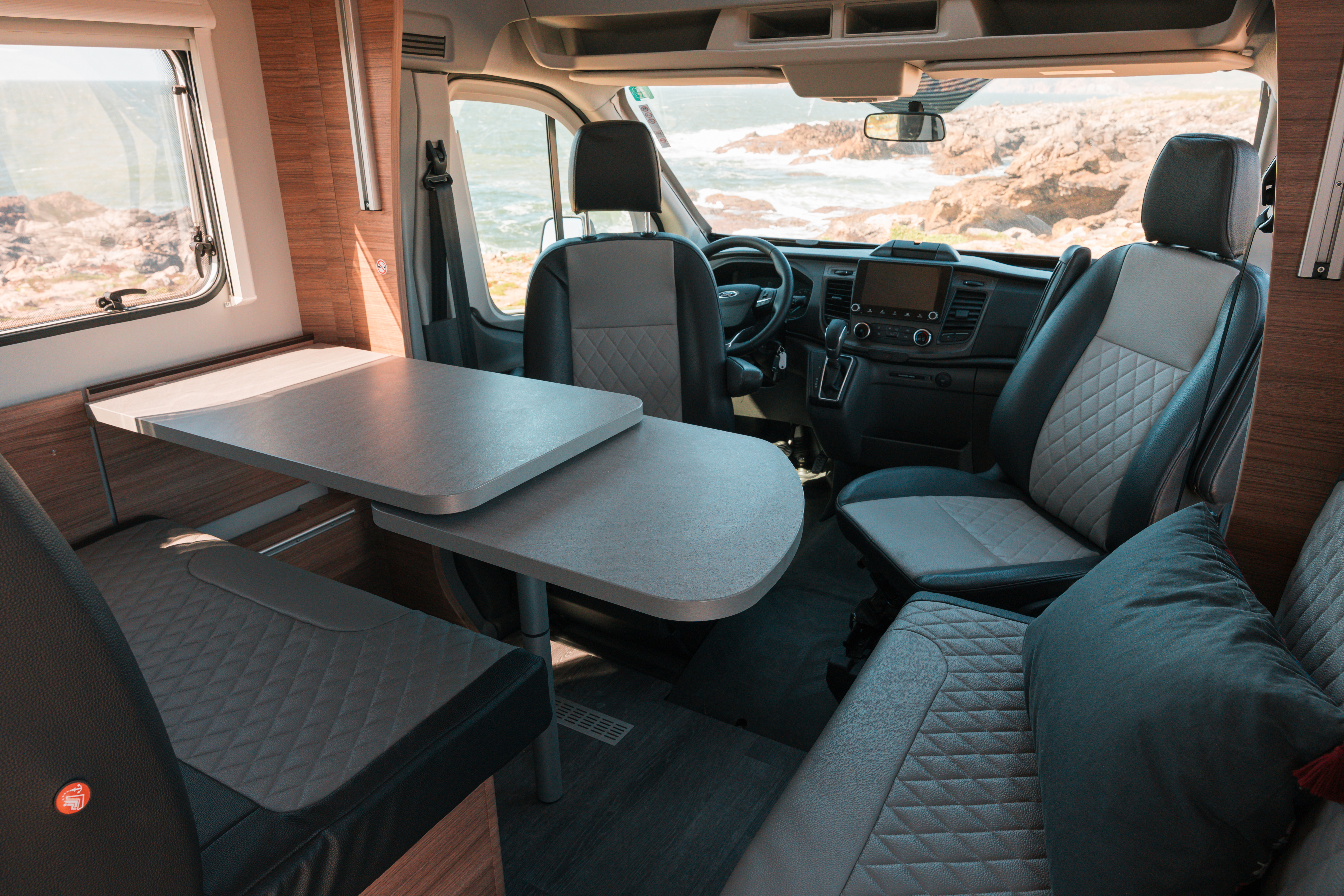 Van interior with front lounge area, swivel seats, dining table, and padded seating, with dashboard and steering wheel visible.