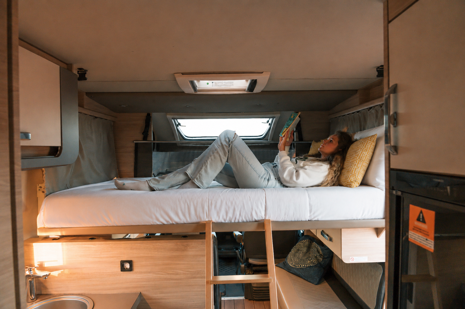 Van interior with upper bed, ladder, storage cabinets, small sink, and reading light visible.