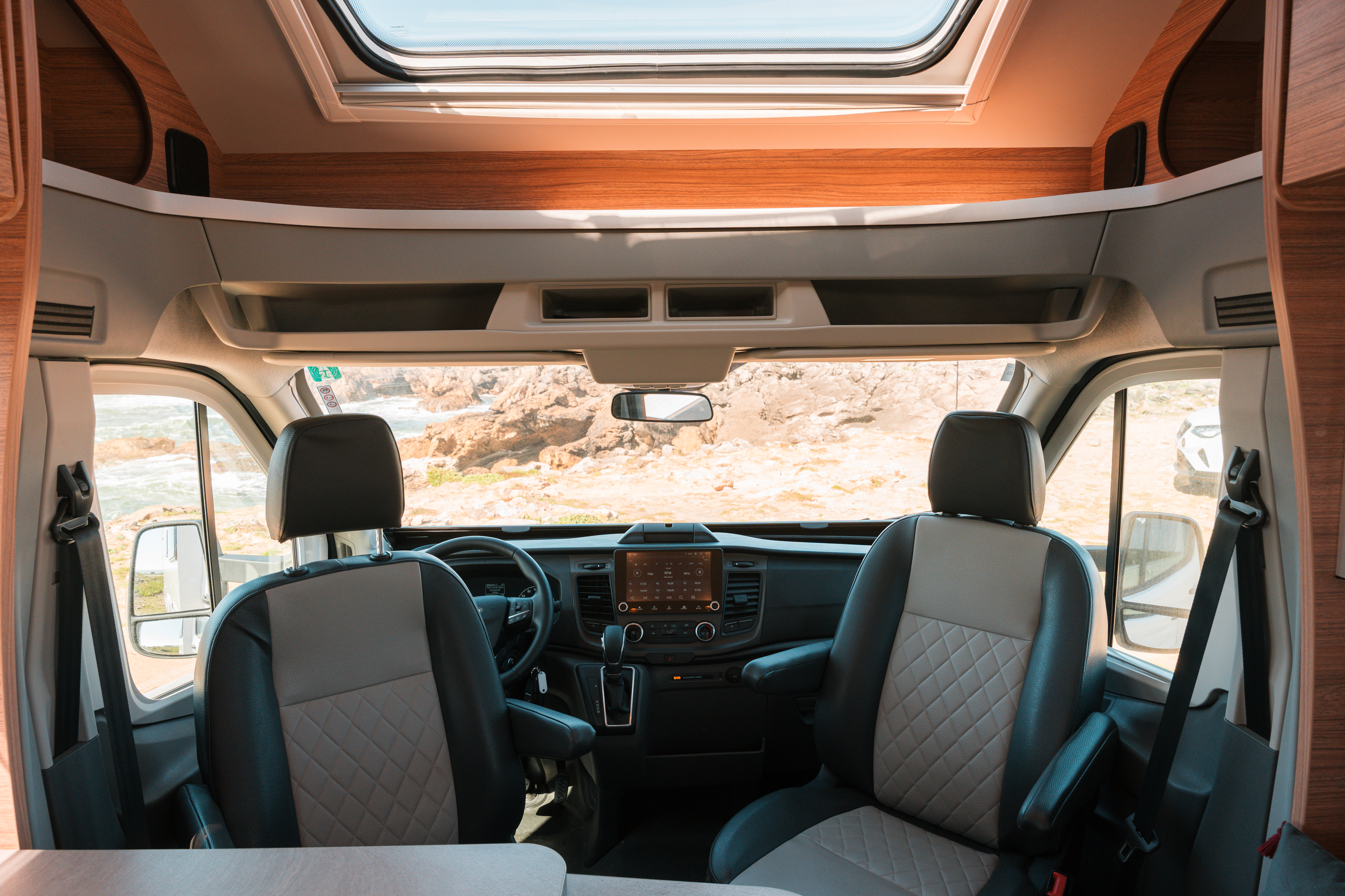 Front camper van interior with two swivel seats, dashboard, overhead storage, and skylight visible.