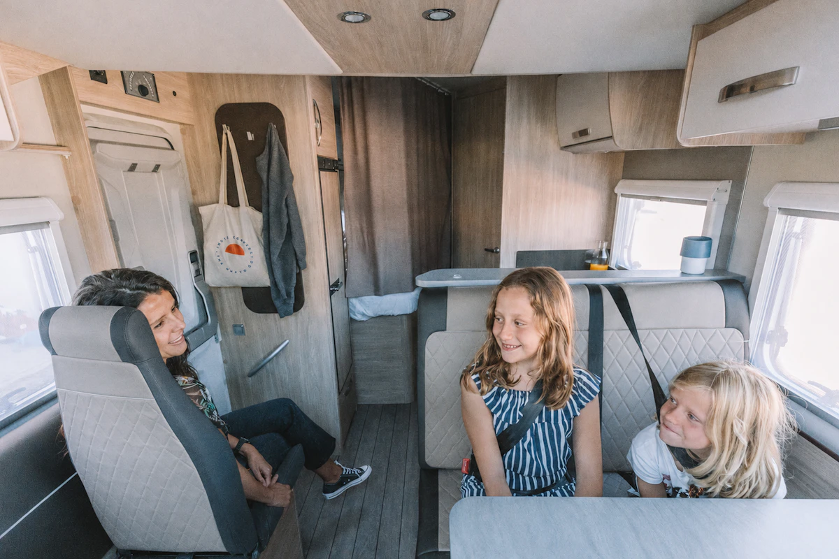 Van interior with seating area, dining table, overhead cabinets, and rear kitchen visible.