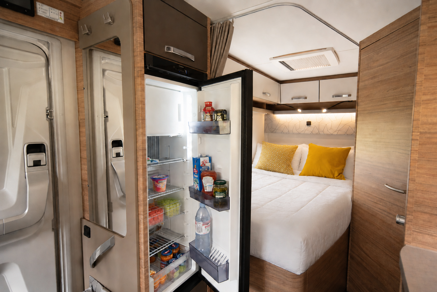Van interior with open fridge, bed with two pillows, overhead cabinets, and wooden paneling.