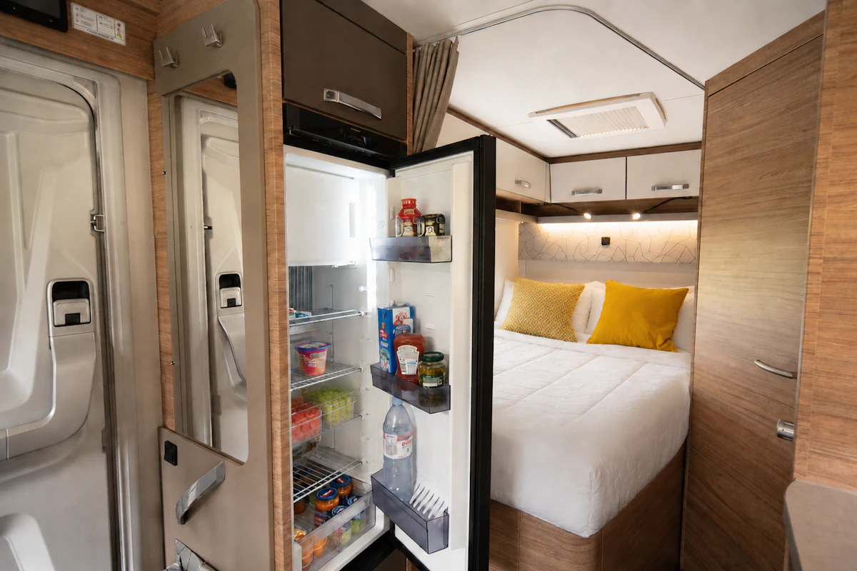 Van interior with open fridge, bed with two pillows, overhead cabinets, and wooden paneling.