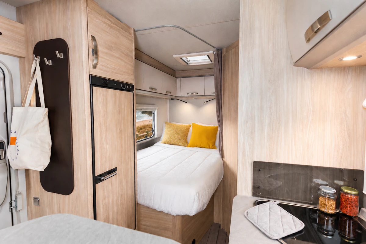Van interior with a rear bed, kitchen area, overhead cabinets, and compact fridge visible.