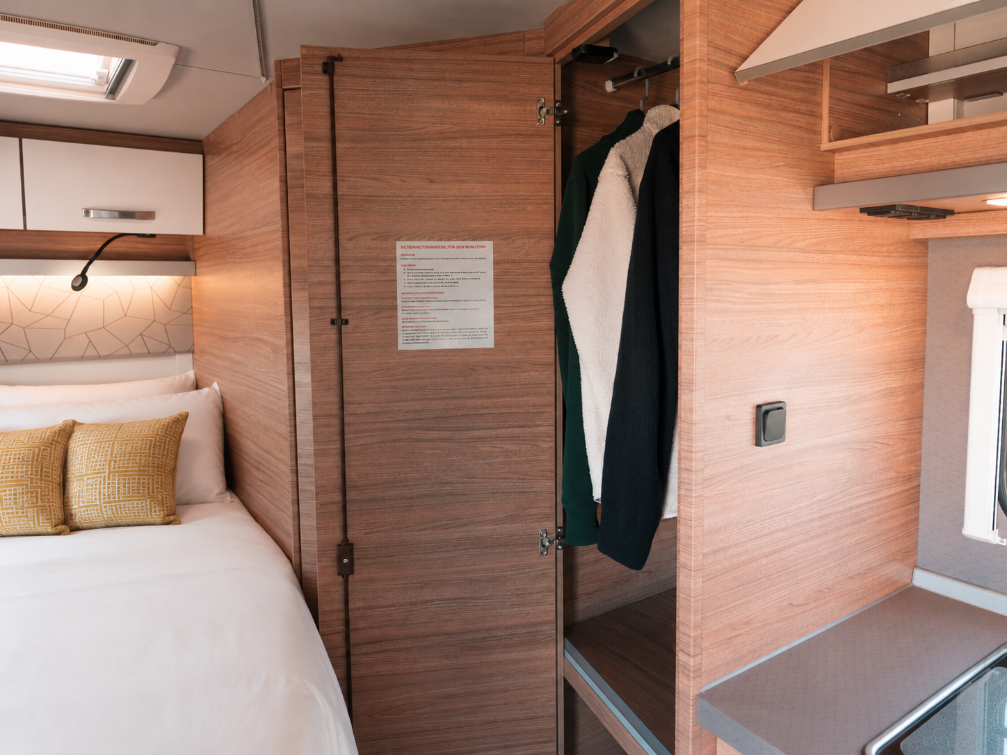 Van interior with bed, pillows, overhead storage, open wardrobe with clothes, wooden paneling, and small counter.