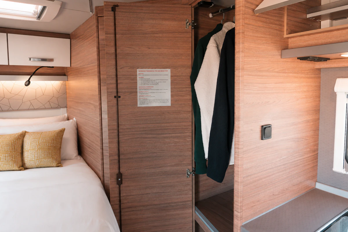 Van interior with bed, pillows, overhead storage, open wardrobe with clothes, wooden paneling, and small counter.