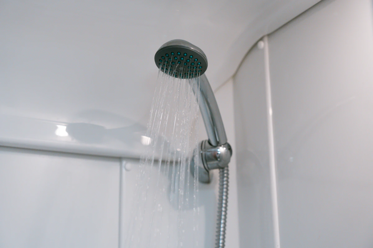 Showerhead with running water in a van bathroom, silver fixtures, white walls and curved ceiling visible.