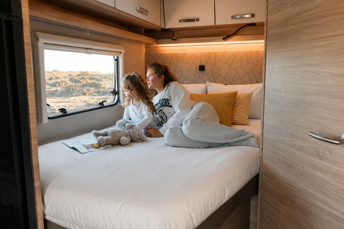 Van interior with large bed, overhead cabinets, decorative pillows, side window, and soft lighting visible.