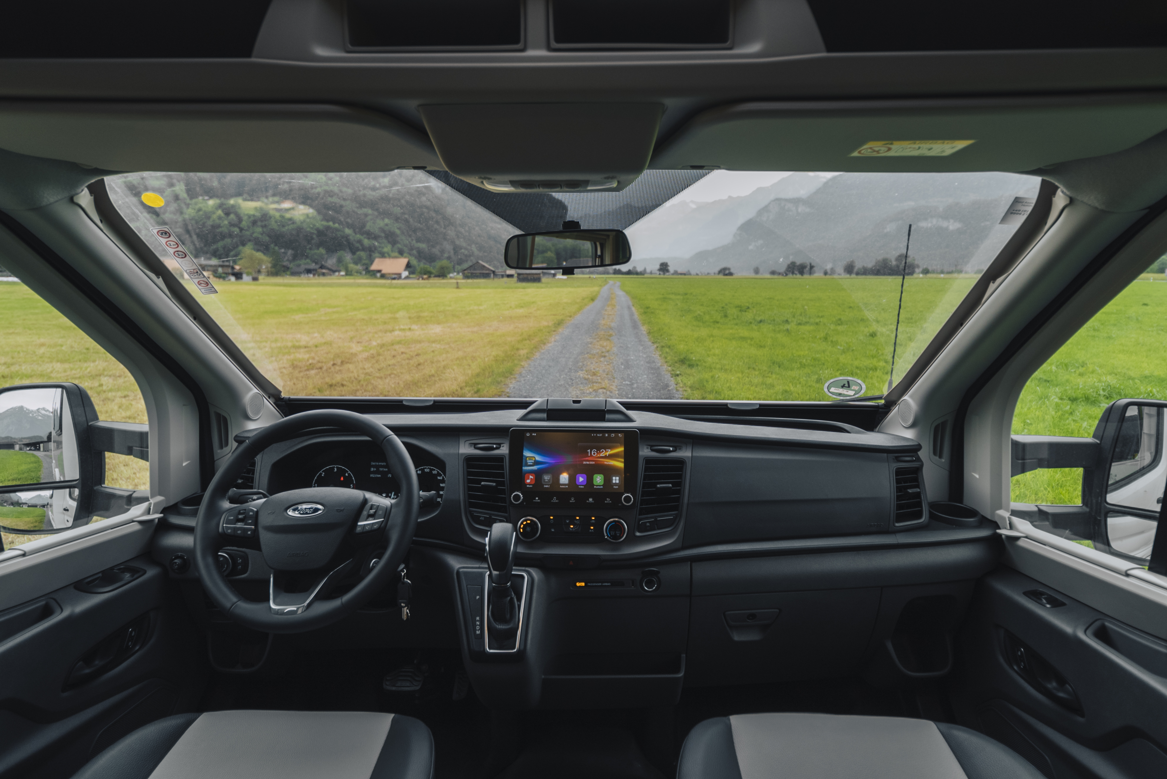 Van interior showing front seats, dashboard with touchscreen, steering wheel, and gear lever, viewed from inside.