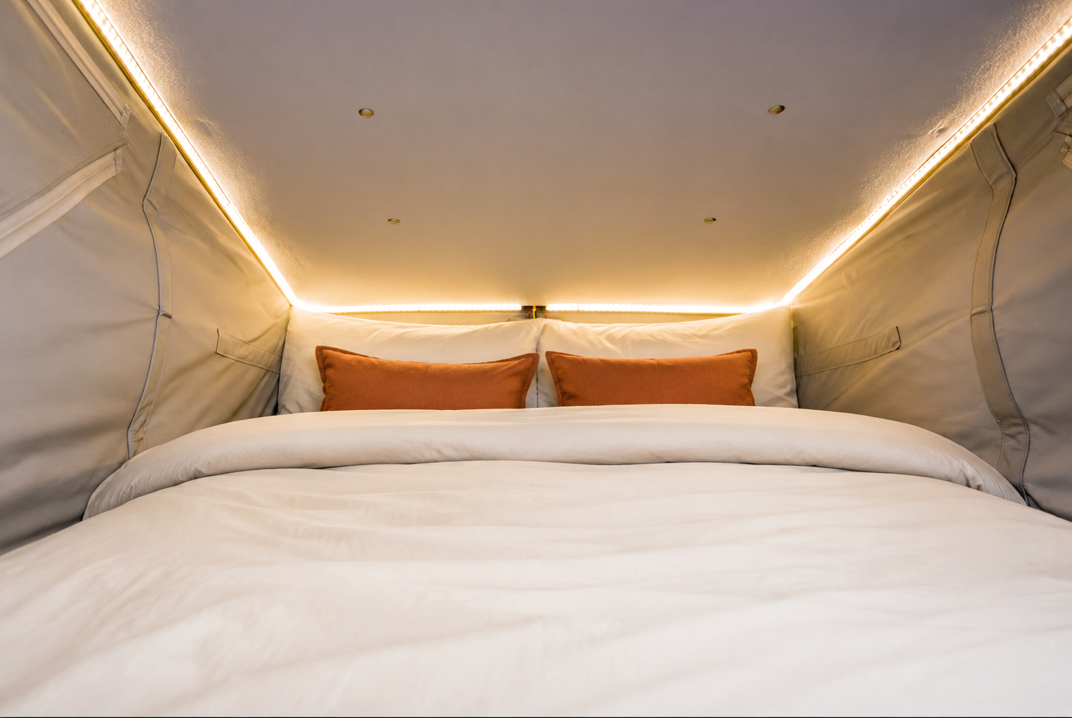 Van interior showing a raised double bed with two pillows, ceiling lights, and fabric side walls.