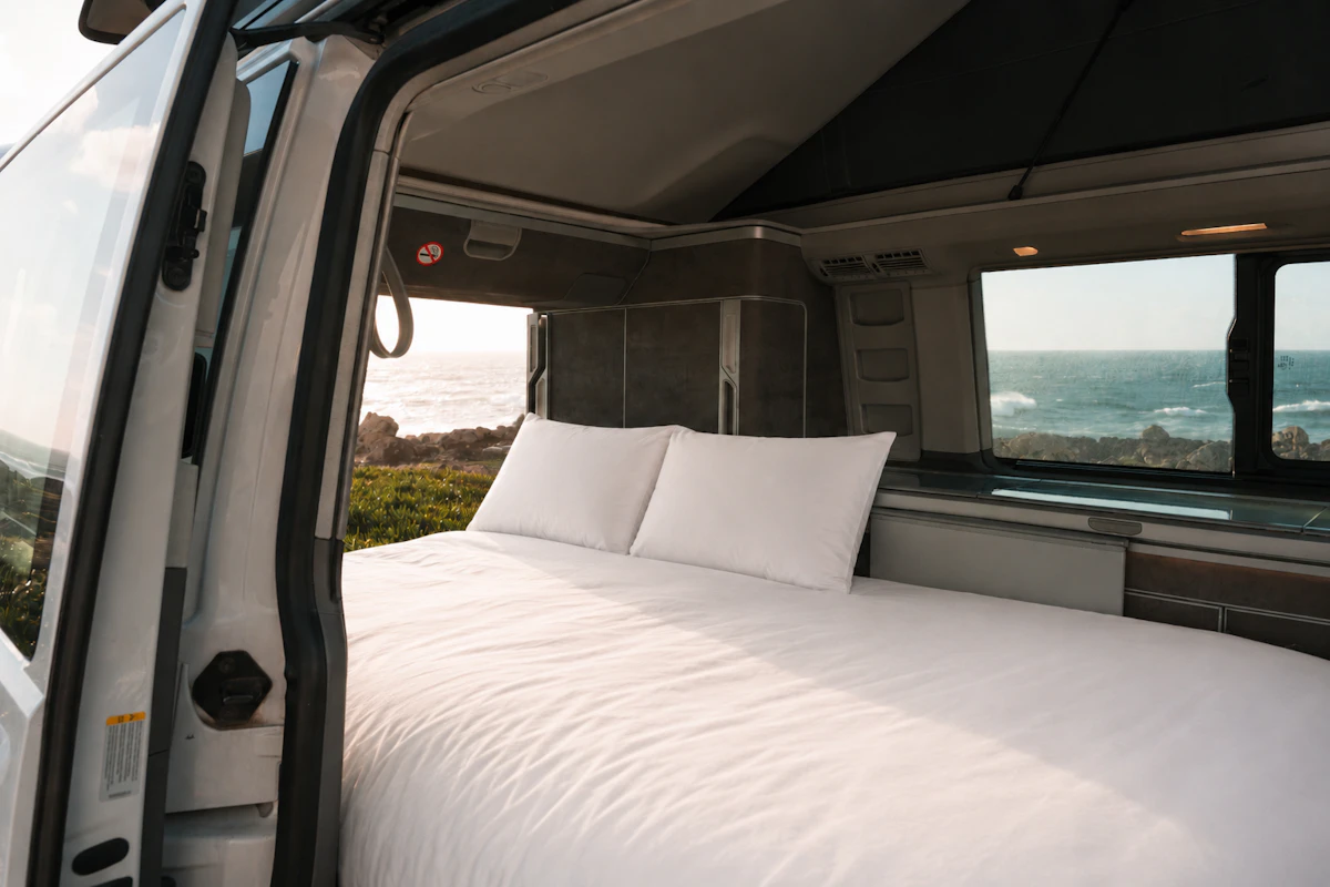 Van interior with made bed, pillows, cabinets, and window, viewed from the open side door.