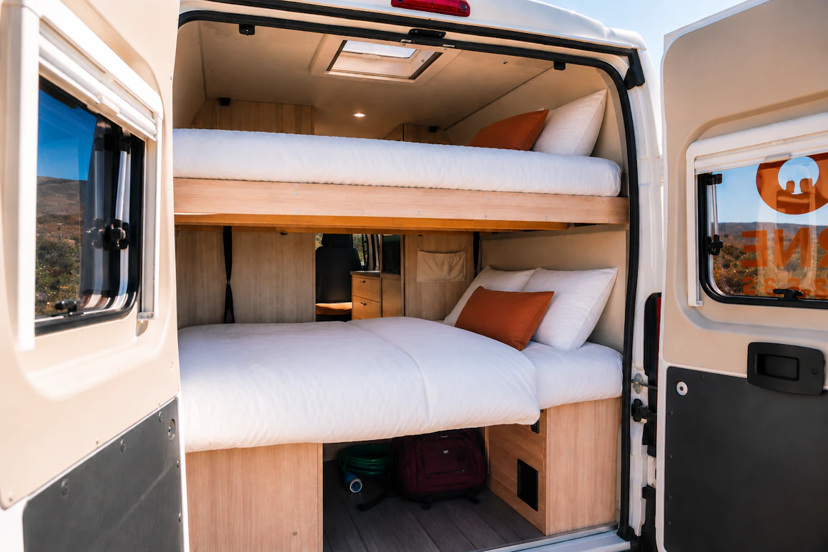 Van interior with two bunk beds, light wood paneling, storage compartments, and visible bedding and pillows.
