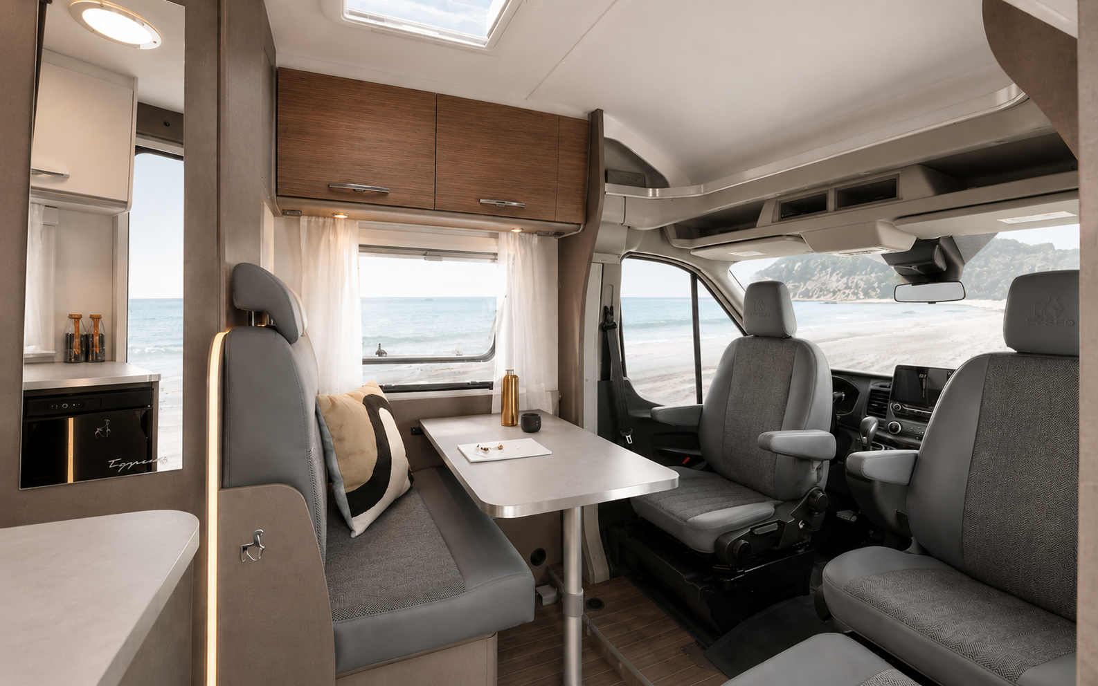 Camper van interior with front seating area, dining table, overhead cabinets, and visible kitchen features.