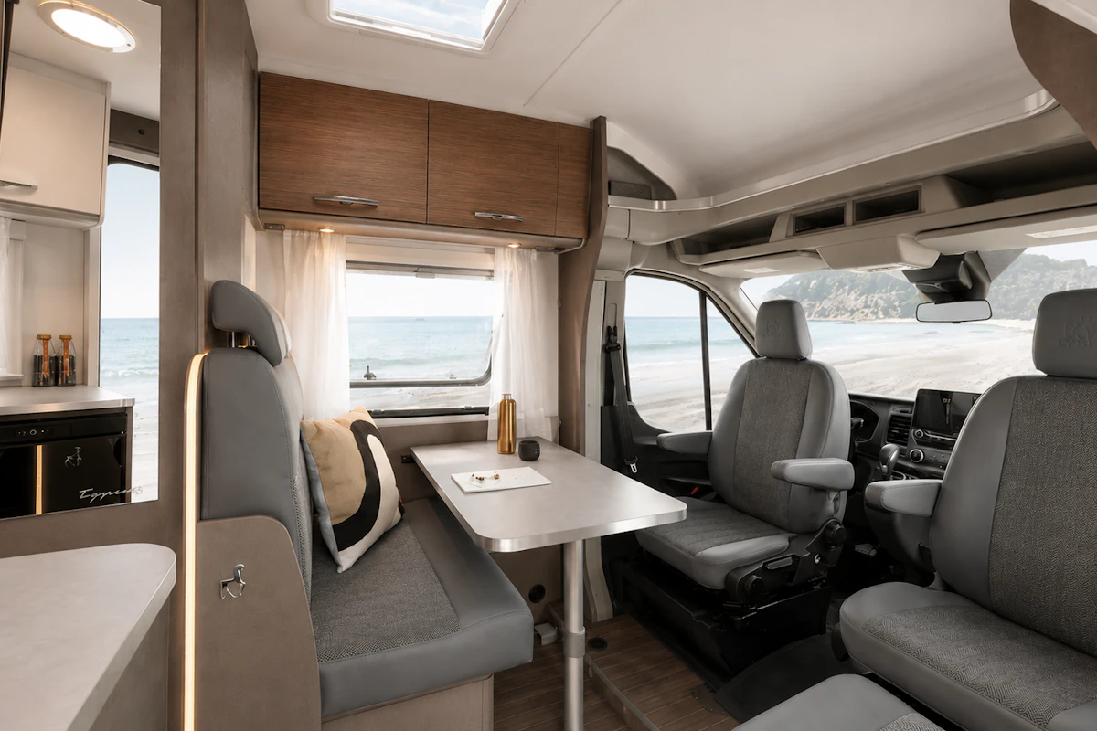 Camper van interior with front seating area, dining table, overhead cabinets, and visible kitchen features.