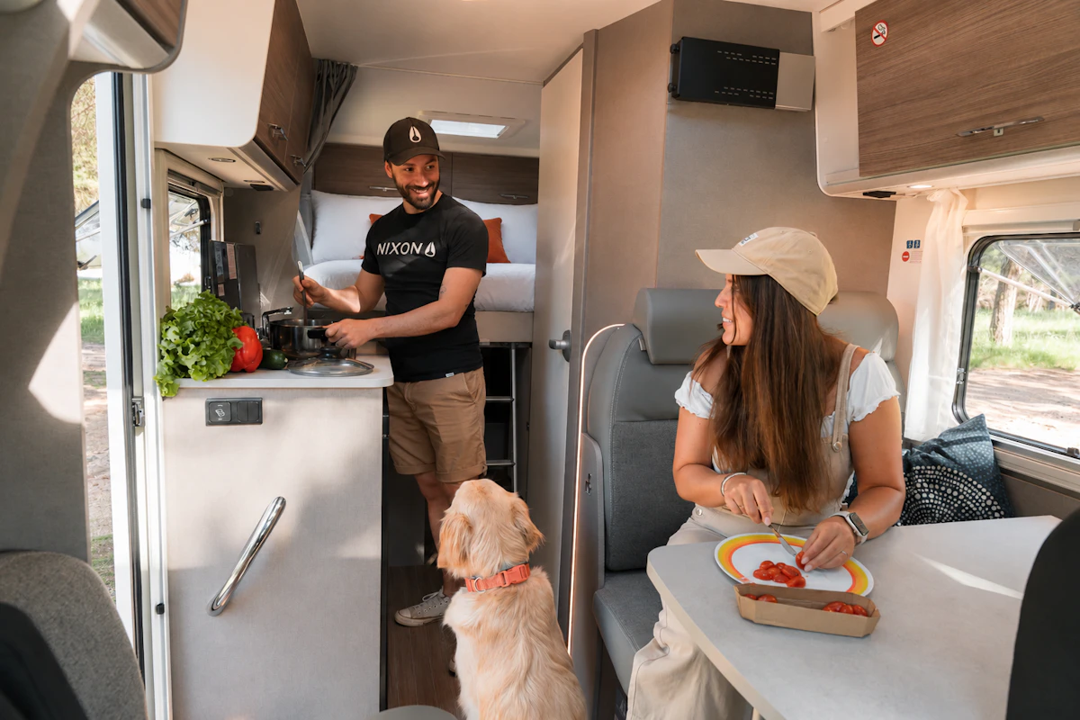 Couple cooking and eating inside a camper van with their dog, fresh vegetables on the counter, bright natural light.