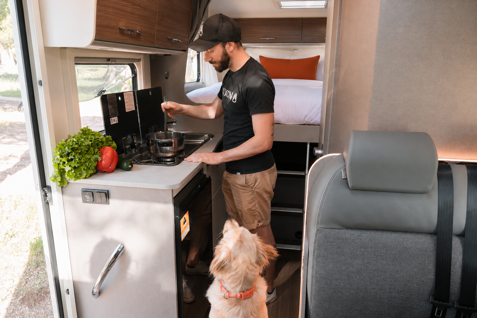 Kitchen area inside camper van with stove, sink, counter, overhead cabinets, and visible fixed rear bed.