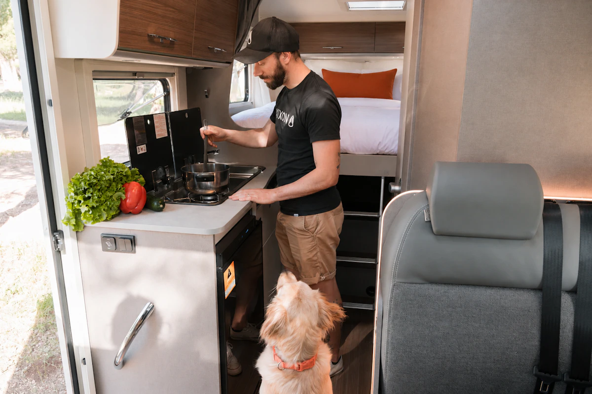 Kitchen area inside camper van with stove, sink, counter, overhead cabinets, and visible fixed rear bed.