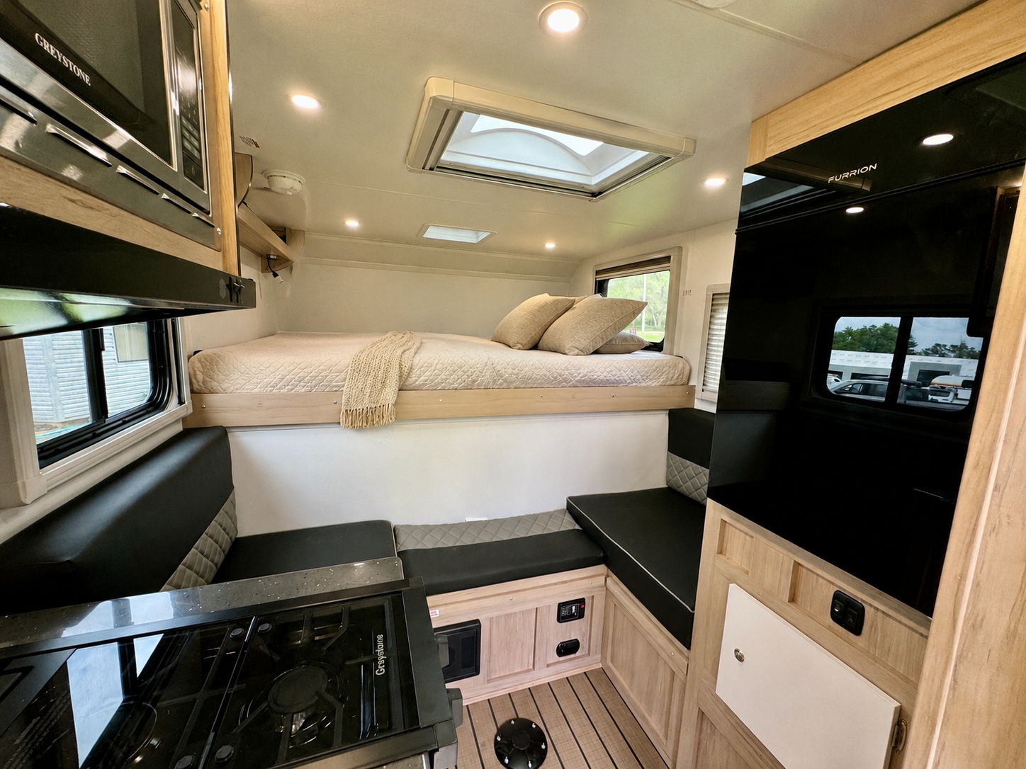 Compact van interior with elevated bed, kitchenette with stove, black benches, storage cabinets, and skylight.