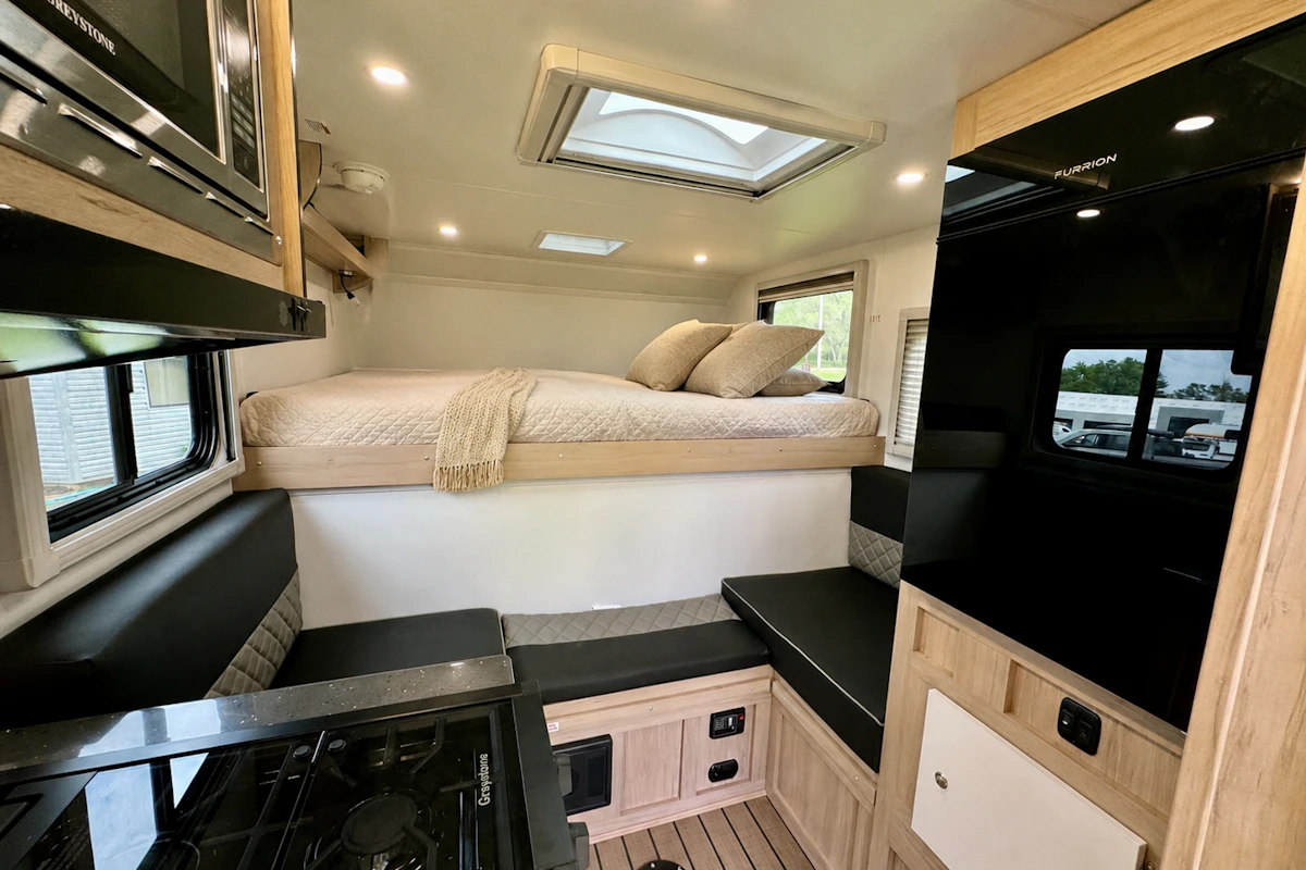 Compact van interior with elevated bed, kitchenette with stove, black benches, storage cabinets, and skylight.