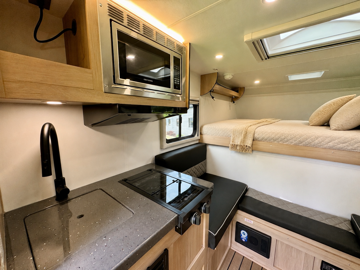 Kitchen area with sink, stove, microwave, seating, and fixed upper bed with pillows and blanket inside the van.