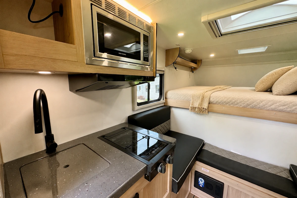 Kitchen area with sink, stove, microwave, seating, and fixed upper bed with pillows and blanket inside the van.