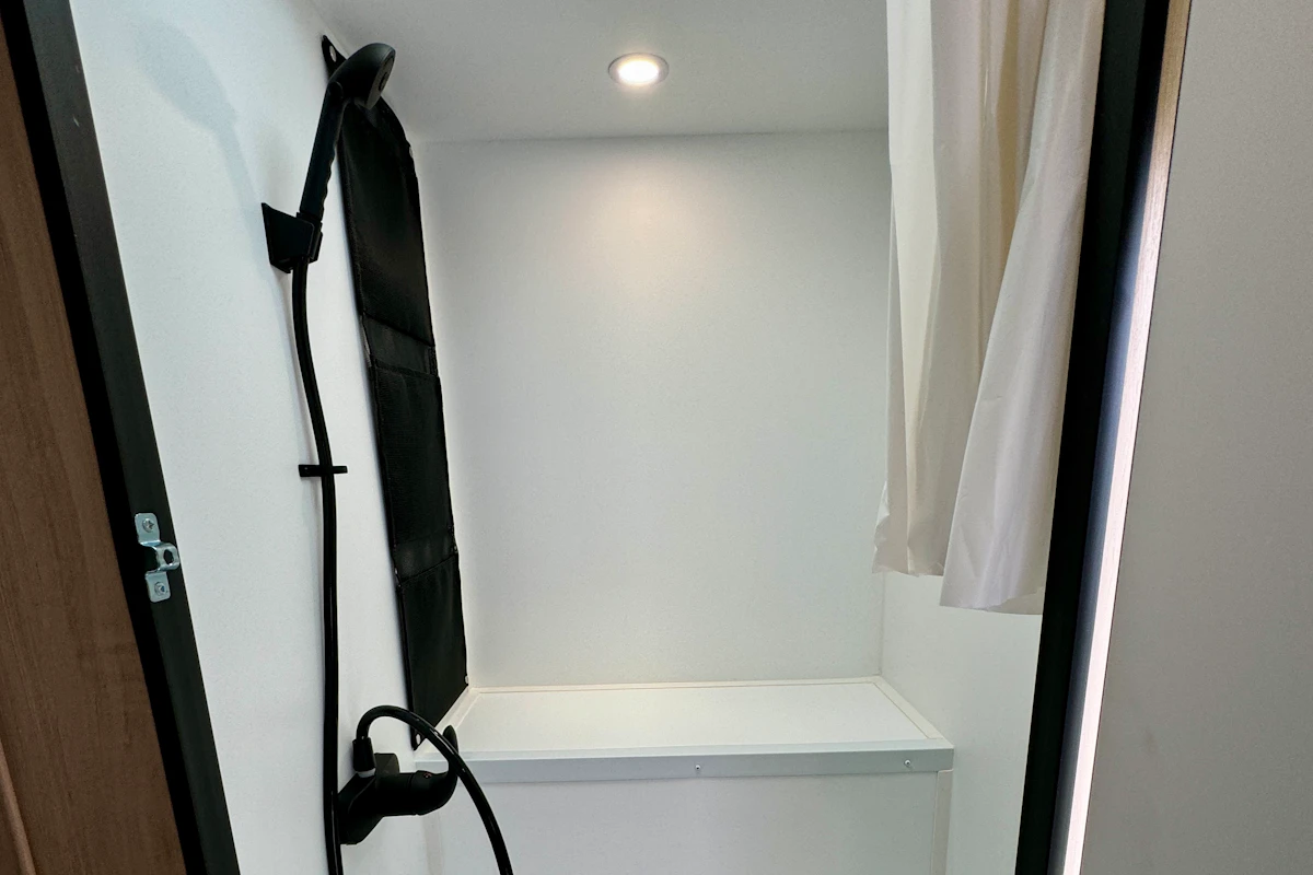 Van interior with shower area, wall-mounted showerhead, white walls, ceiling light, and partial curtain visible.