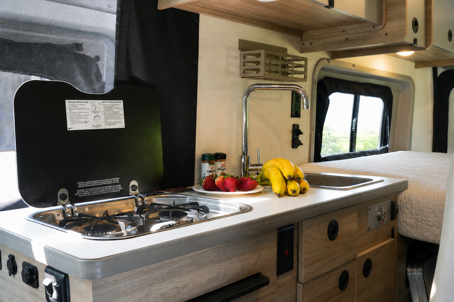 Camper van interior kitchen with stove, sink, countertop, storage drawers, and part of the bed visible.