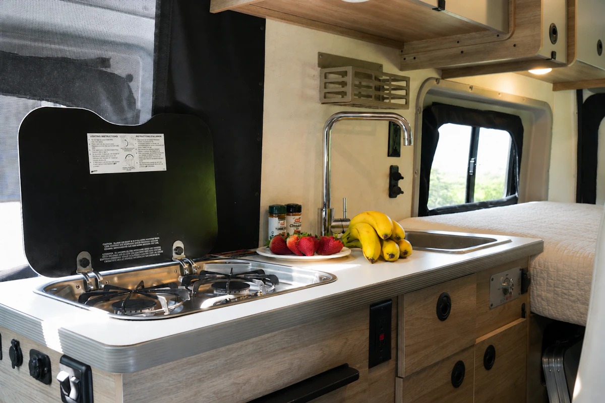 Camper van interior kitchen with stove, sink, countertop, storage drawers, and part of the bed visible.