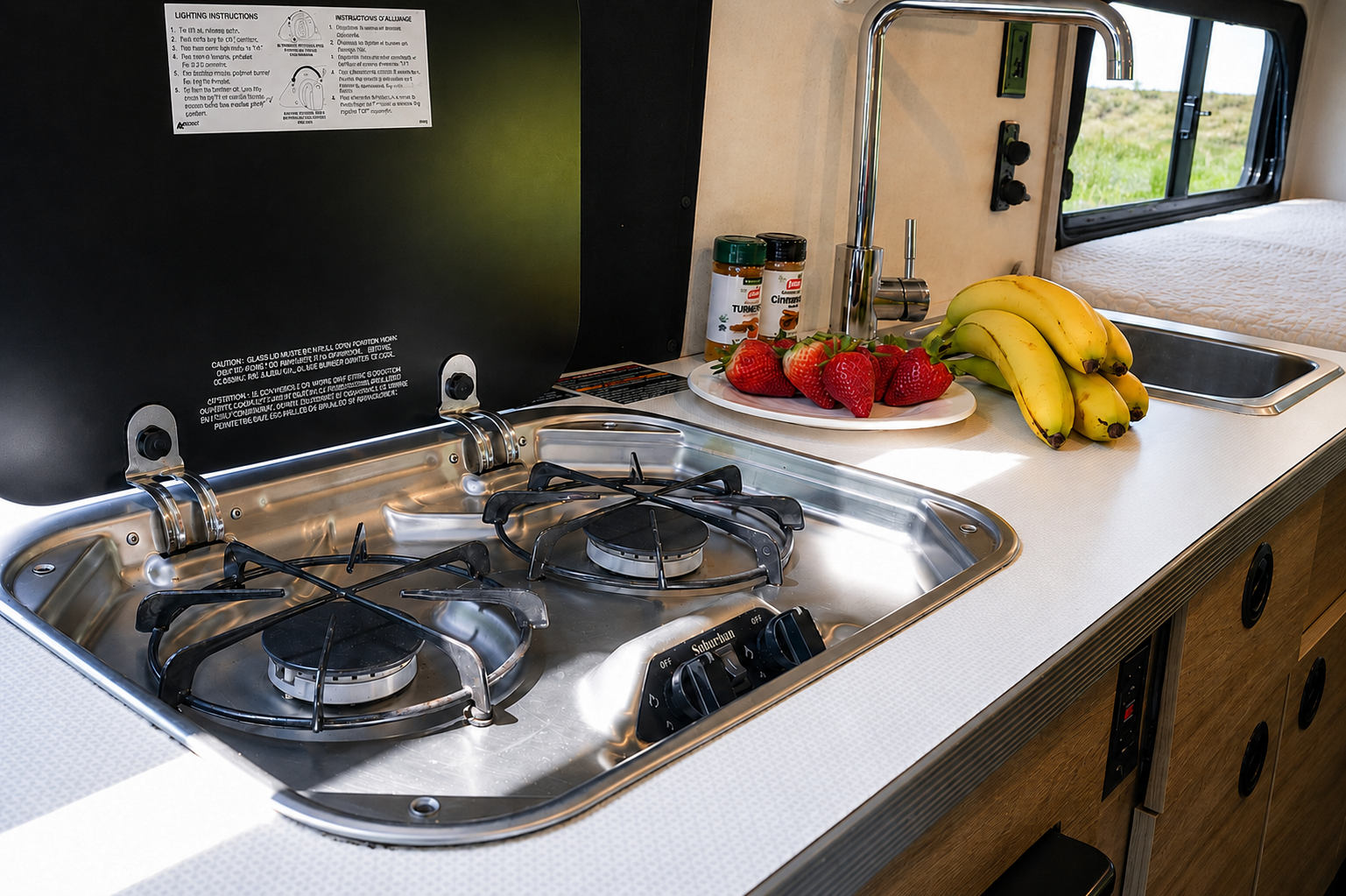 Van interior with two-burner stove, sink, countertop, food items, and storage drawers visible.