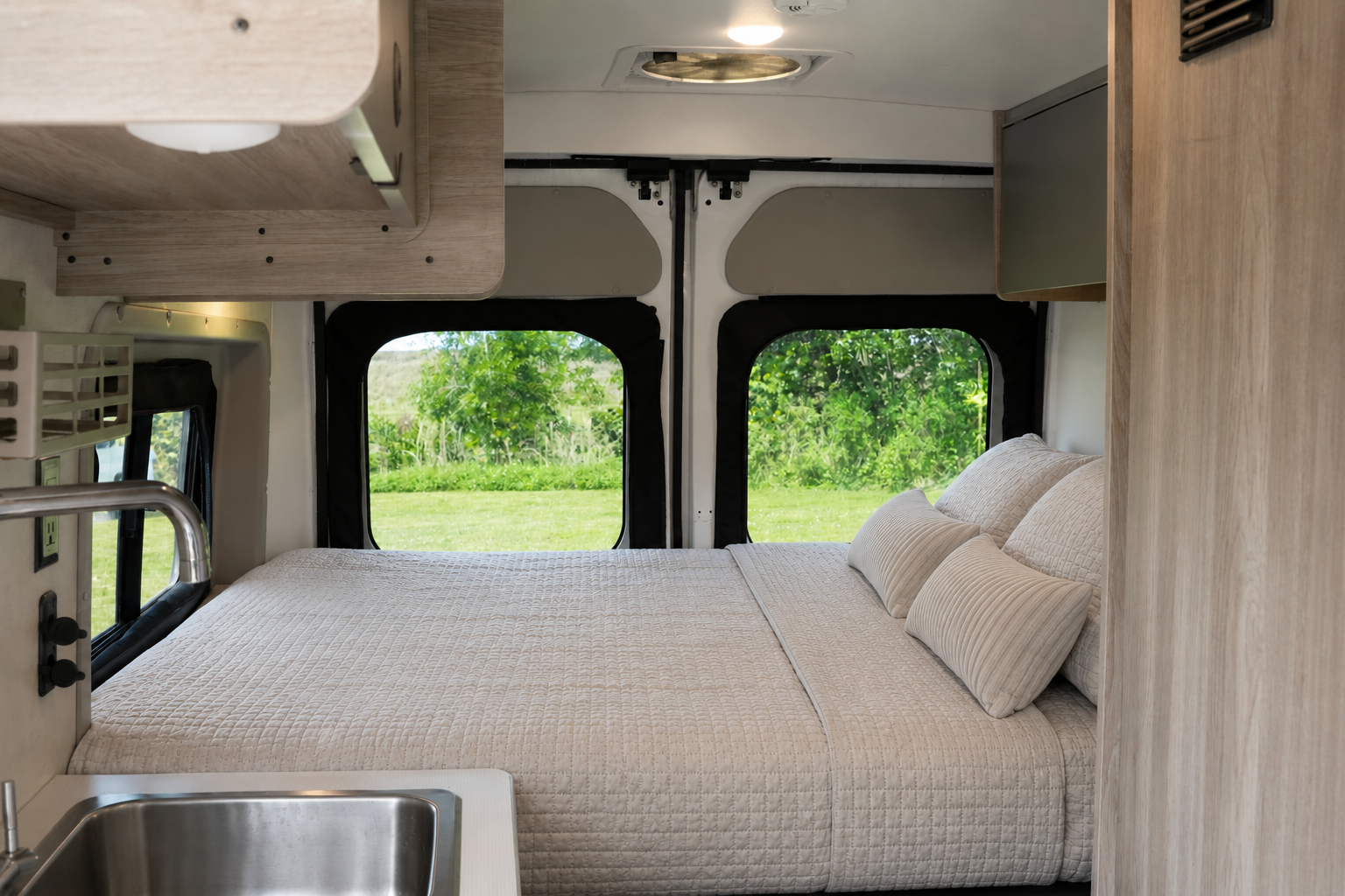 Van interior with large bed, pillows, overhead storage, sink, and windows at the rear.