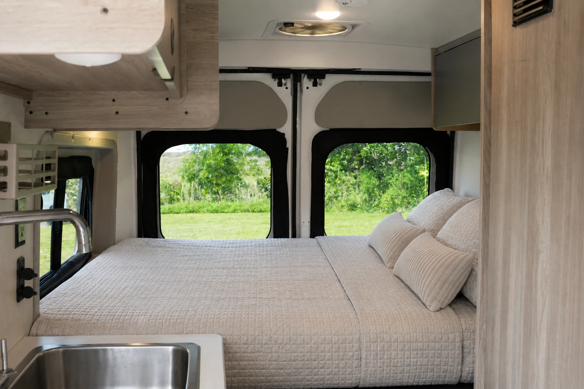 Van interior with large bed, pillows, overhead storage, sink, and windows at the rear.