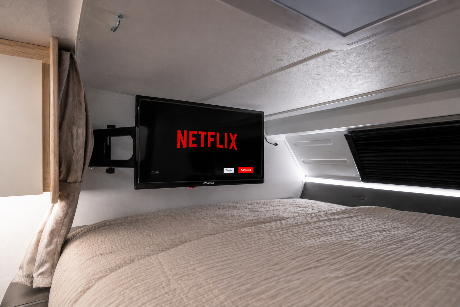Bed area with mounted TV screen, light bedding, cabinet, window, and curtain inside camper van.