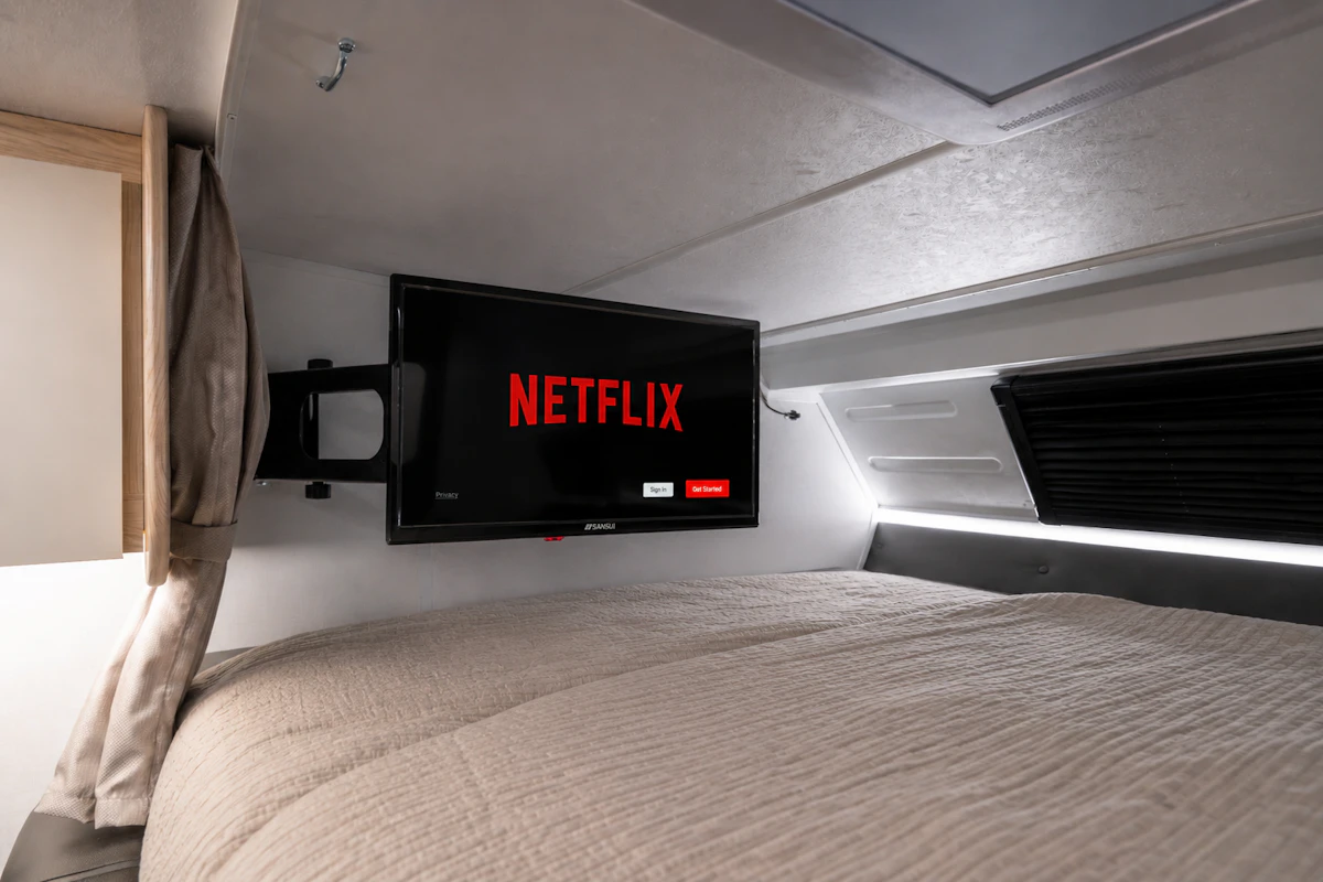 Bed area with mounted TV screen, light bedding, cabinet, window, and curtain inside camper van.