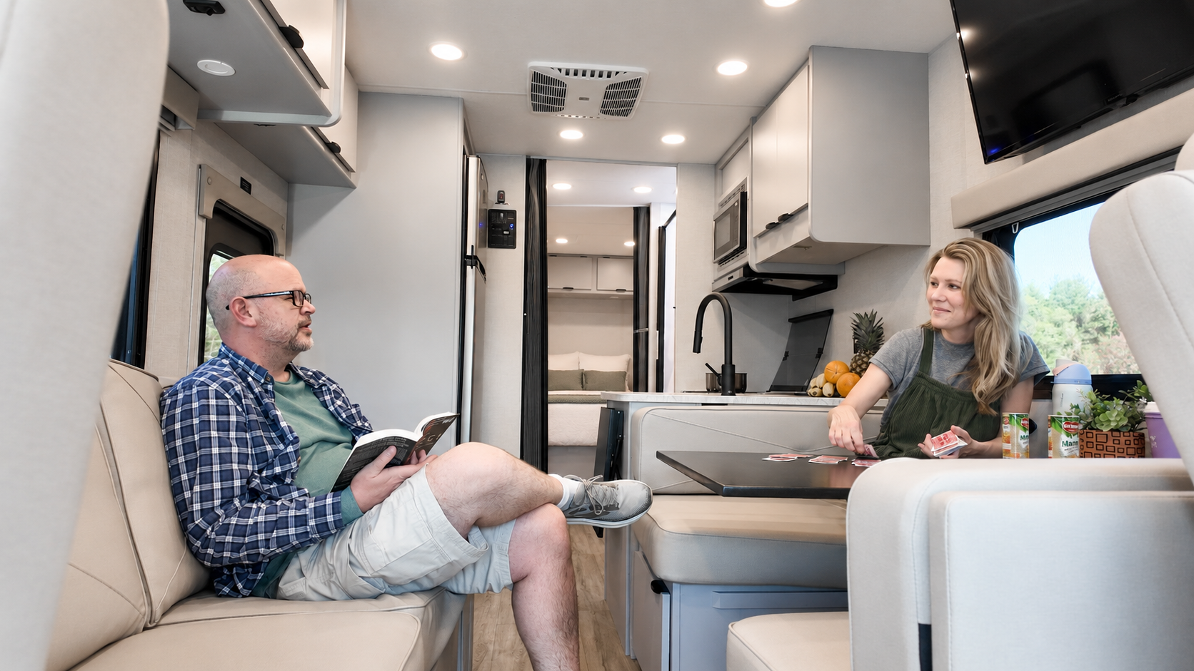 Camper van interior with seating area, table, kitchen, overhead cabinets, and bed visible in the background.
