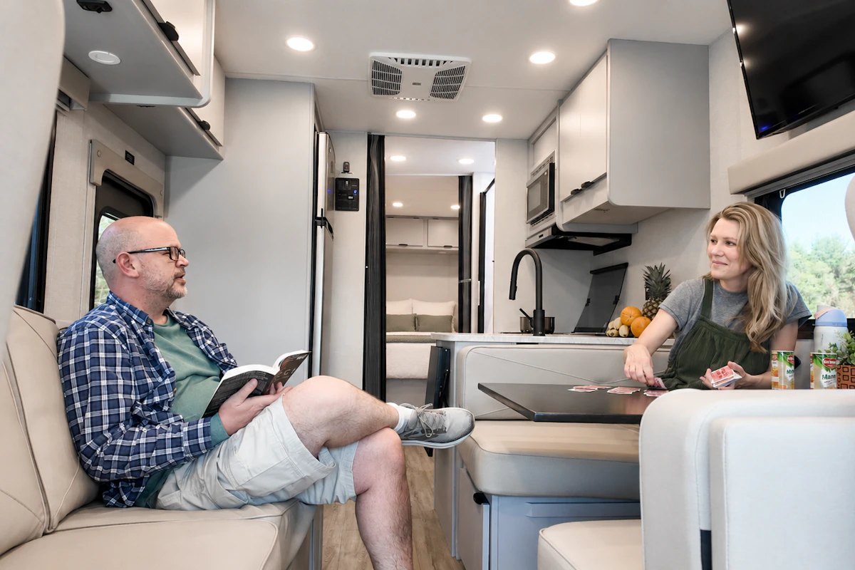 Camper van interior with seating area, table, kitchen, overhead cabinets, and bed visible in the background.
