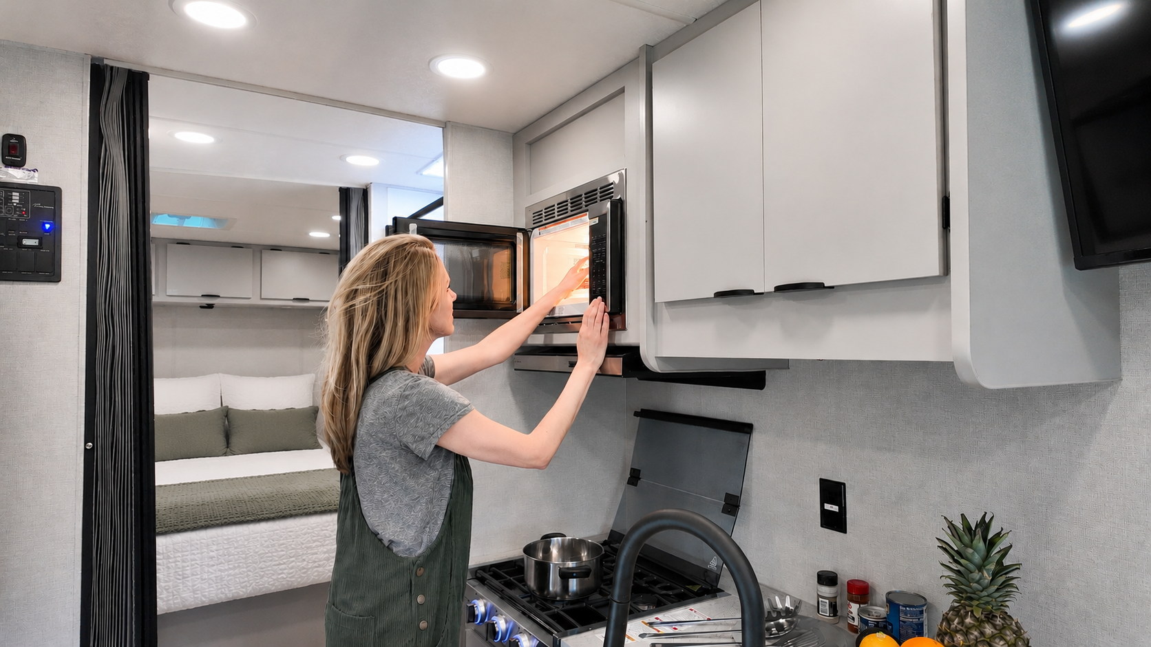Compact van kitchen interior with microwave, stove, sink, cabinets, and visible bed area in the background.