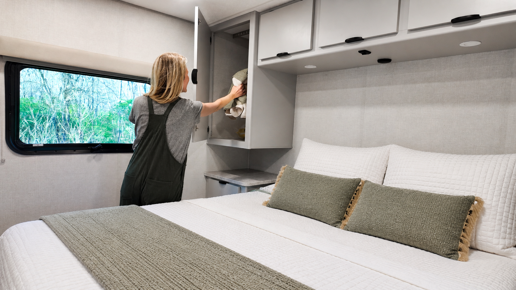 Van interior with bed, overhead cabinets, window, and person opening a storage compartment above a side table.