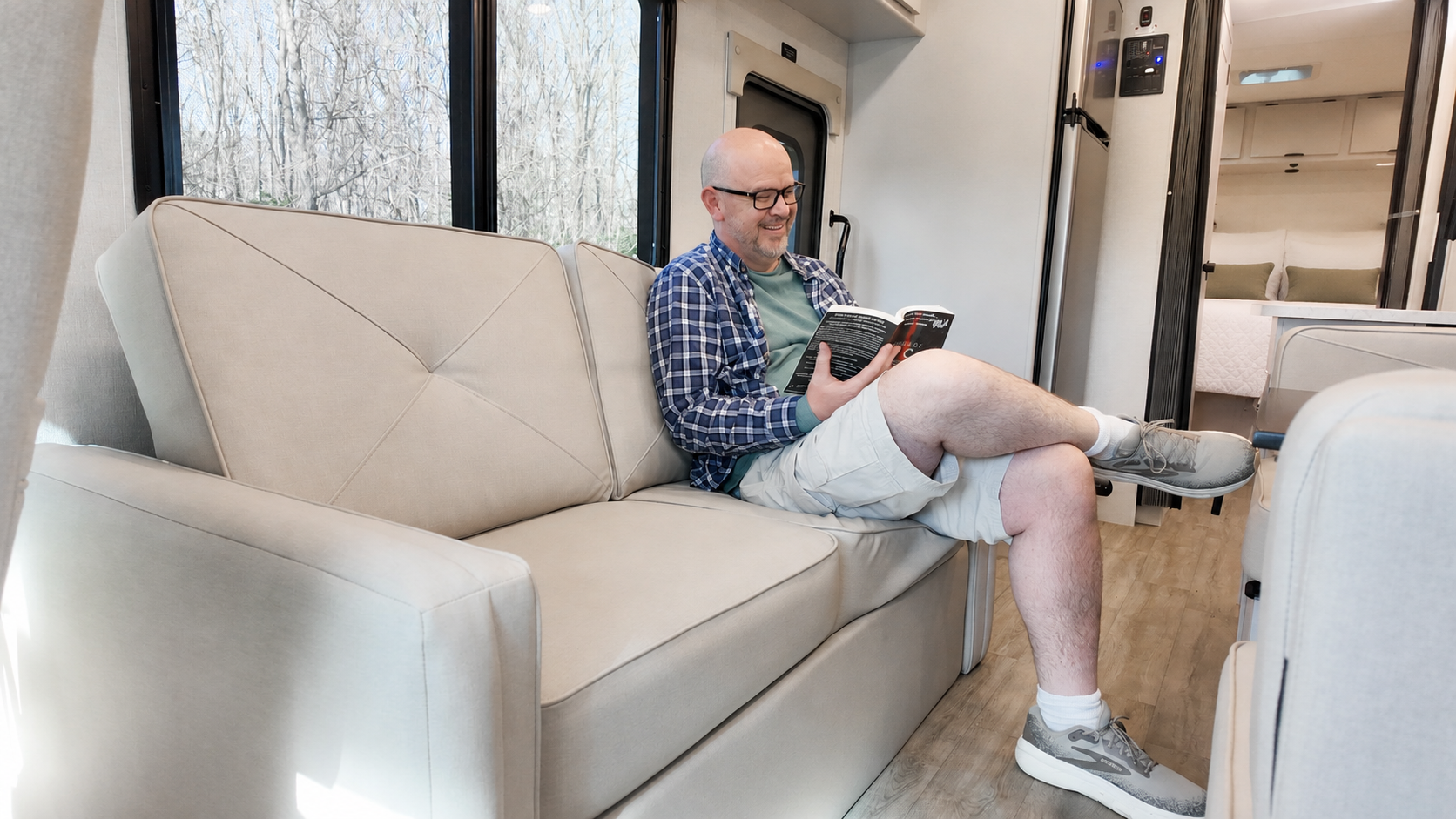 Van interior with beige sofa, reading area, wood flooring, visible kitchenette, and sleeping area in the background.
