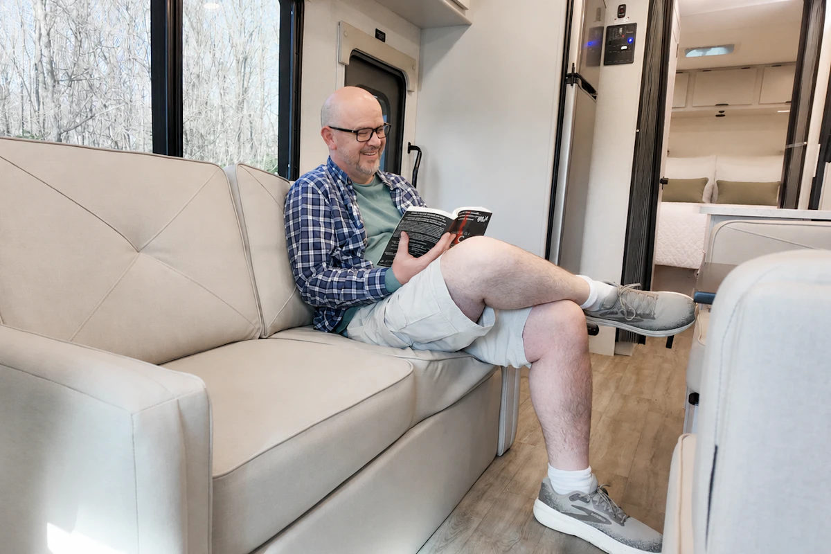 Van interior with beige sofa, reading area, wood flooring, visible kitchenette, and sleeping area in the background.