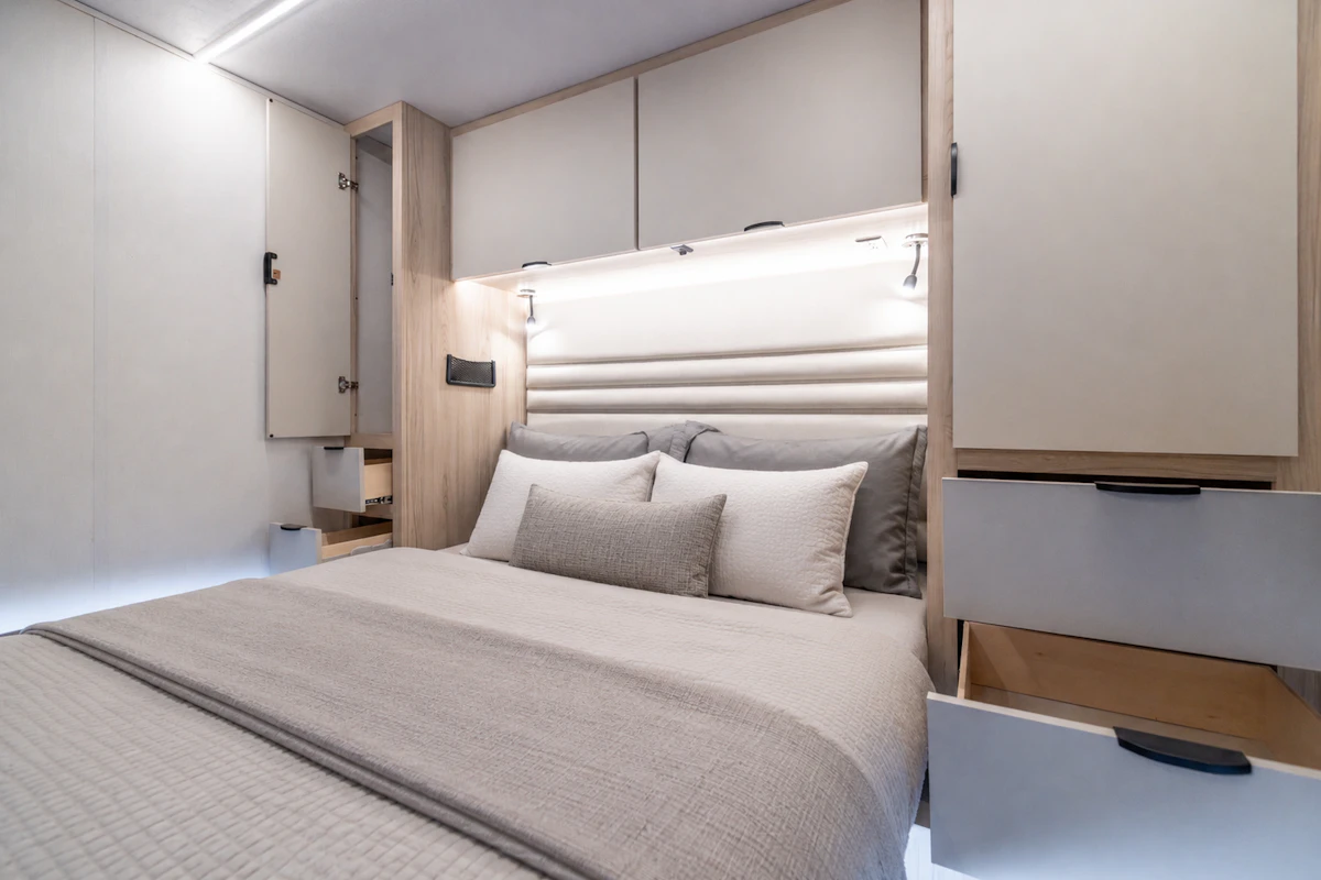 Double bed with pillows, overhead cabinets, open drawers, and wall lights inside camper van bedroom.