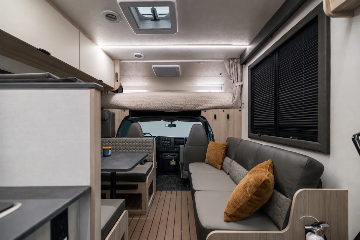 Modern camper van interior with sofa, dining table, cabinets, and blinds, viewed from the rear toward the driver's cabin.