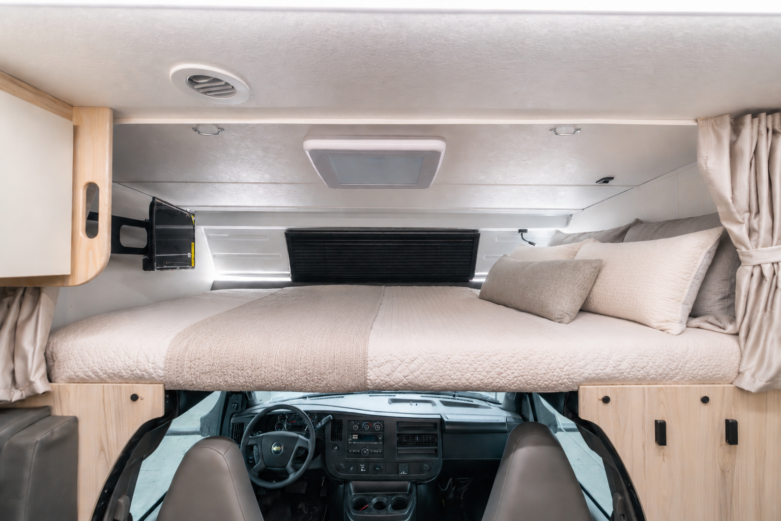 Over-cab bed area above front seats with bedding, pillows, side cabinets, small TV, and curtain dividers.